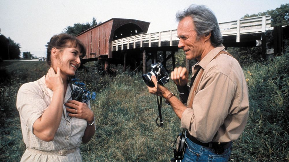 The Bridges of Madison County. Meryl Streep and Clint Eastwood look lovingly at each other as he photographs her in grass beside a bridge.