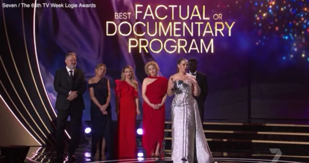 A woman in a silver dress is flanked by four others accepting an award for best factual or documentary program.