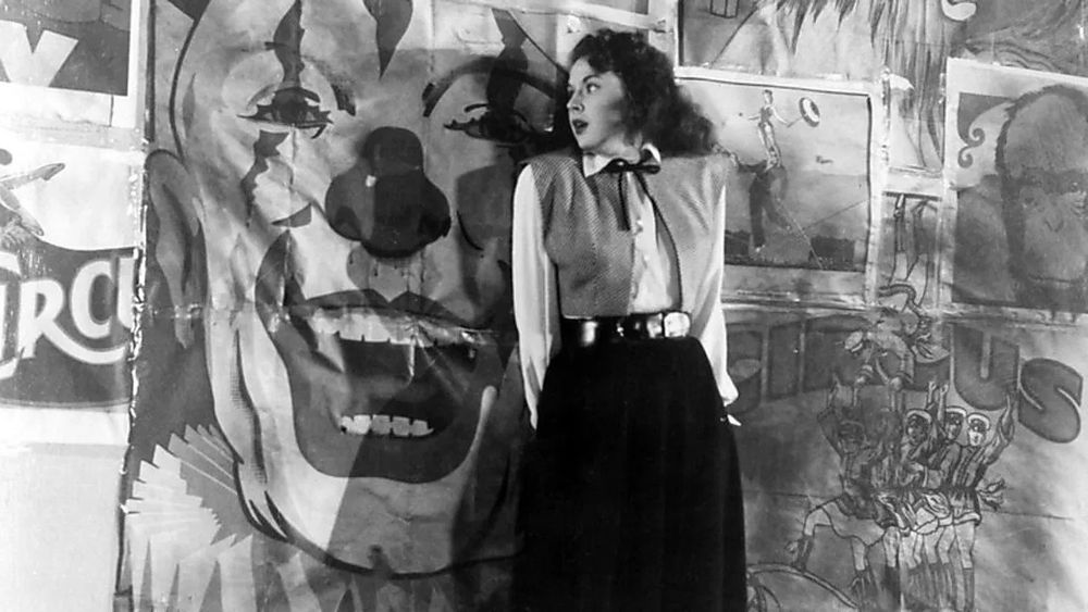 Outrage. A young woman is scared on a street next to a large scary clown poster.
