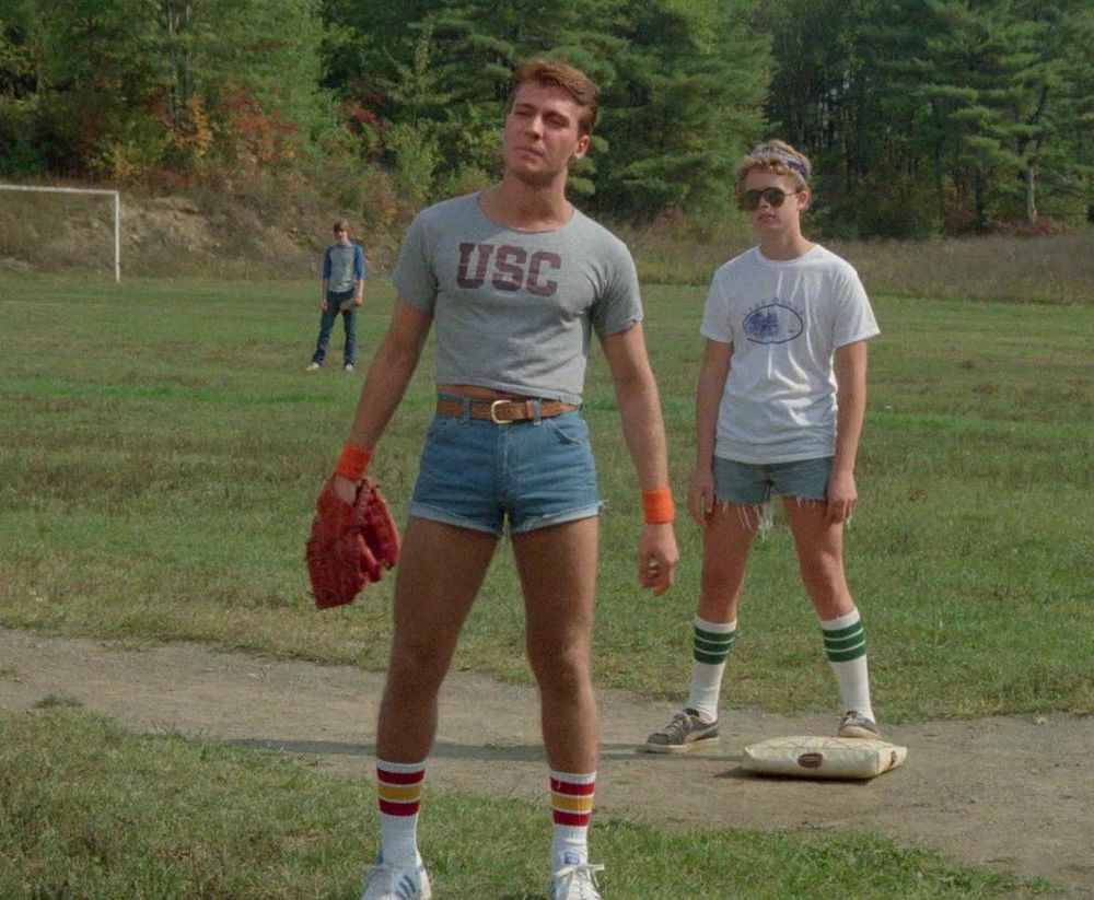 A man wearing a crop shirt and belted denim shorts and catchers mitt on a camp baseball field.