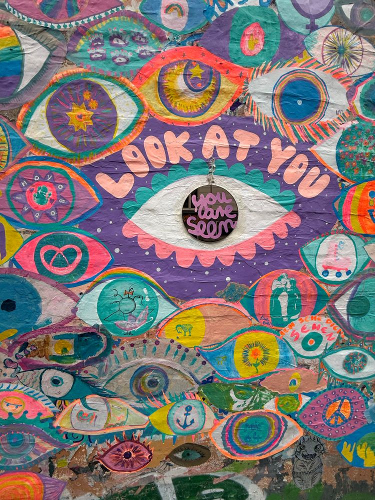 Eine bunt gestaltete Häuserwand mit gemischten Streetart-Elementen. Viele phantasievolle Augen sehen den Betrachtenden. Mittig ein größeres Auge mit Spiegel als Pupille, dazu der Text „Look at you“ und „You are seen“. Künstler*in unbekannt.