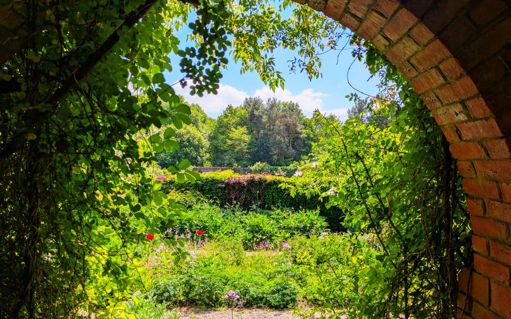 In the foreground close to the camera is an arch, the right side is an arch of bricks, the left side has a trestle covered in vines. Through the arch gardens can be seen, with rows of small plants and flowers separated by gravel paths and hedges. In the distance a line of trees demarcate the start of a woods. Above the tree line a pale blues sky contains bright white low clouds that almost look like the tops of distant mountains.