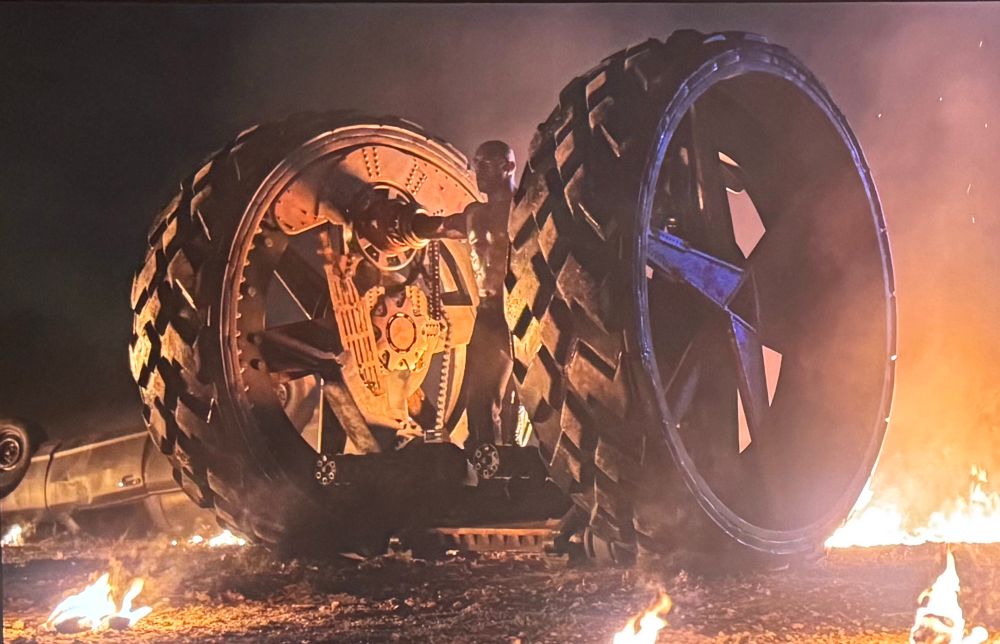 A guy from the show twisted metal who’s arms are inside of giant wheels on either side of him while he stands on a platform in between looking like a huge motorcycle 