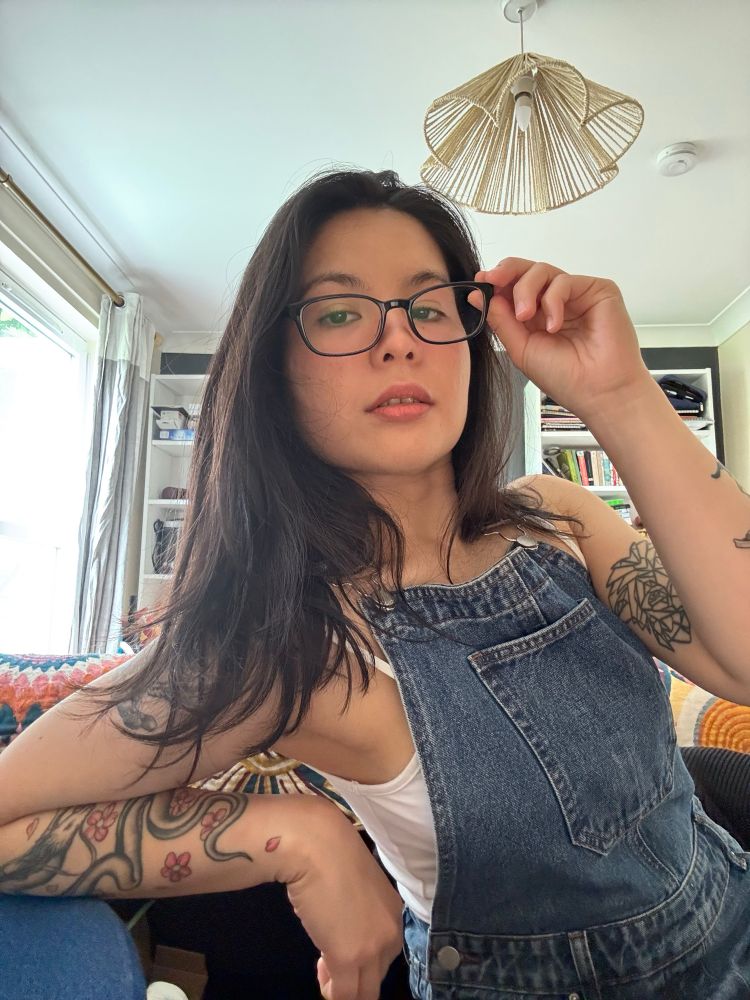 Unsmiling selfie of me in denim dungarees with a white, thin-strapped top underneath. I’m wearing dark-framed rectangular glasses, which I’m holding slightly away from my face