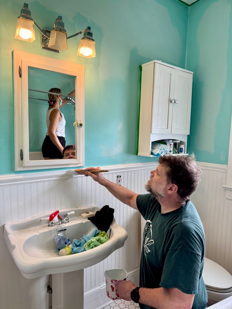 A man (my husband) paints beadboard in a white and aqua bathroom. My friend Jess can be seen painting a wall in the mirror. 