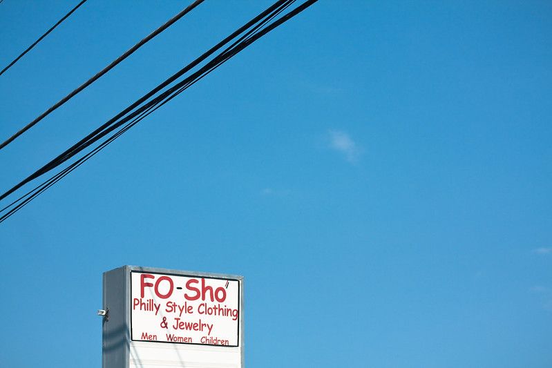Powerlines cross over the top left corner of the frame. A sign says, "Fo Sho Philly Style Clothing and Jewelry."