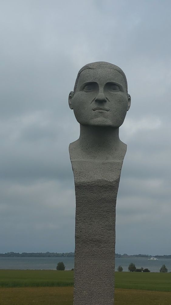 Das Bild zeigt eine markante Steinskulptur, die einen stilisierten menschlichen Kopf darstellt. Die Skulptur wirkt monumental und ist auf einem hohen, rechteckigen Sockel aus demselben Steinmaterial gearbeitet, das sich nach oben hin verjüngt und schließlich in den Kopf übergeht. Der Gesichtsausdruck ist ernst und ruhig, mit symmetrischen Zügen, betonten Augenbrauen und einem leicht verschlossenen Mund.

Im Hintergrund ist eine weite Landschaft mit grünem Gras und einem Gewässer zu sehen – vermutlich ein See oder ein Fjord. Dahinter erstreckt sich eine flache Uferlandschaft mit Bäumen und vereinzelten Gebäuden