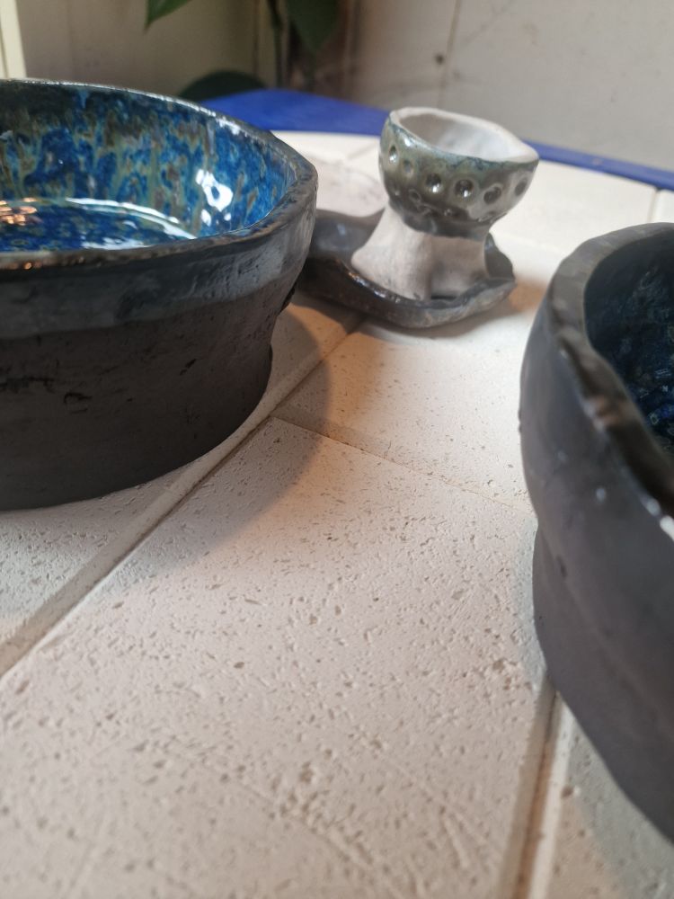 A blue glaze flecked with brown covers the inside of a chunky black stoneware bowl. We can see the side of another and a little egg cup behind. 