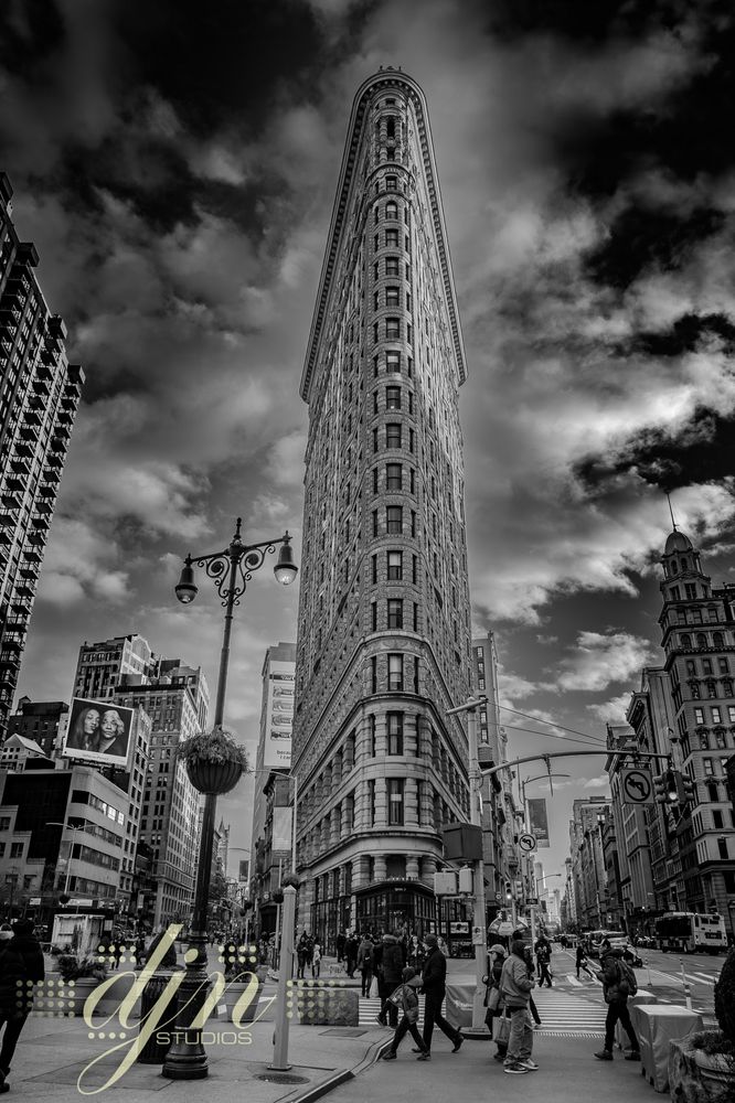 A black and white photo of the Flatiron Building in New York City. The building looks like a fine blade cut against a dramatically clouded sky. Pedestrians travel in every direction across the busy Manhattan streets. 