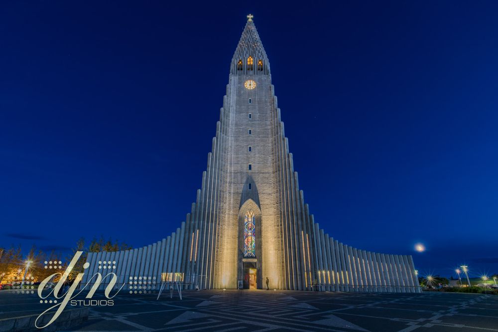 Hallgrímskirkja in Reykjavik. The photo of the church taken at night. The sky is a deep blue. The lights on the front of the church beam yellow, the yellow cast across the front of the church contrast against the blue backdrop of clear sky. 