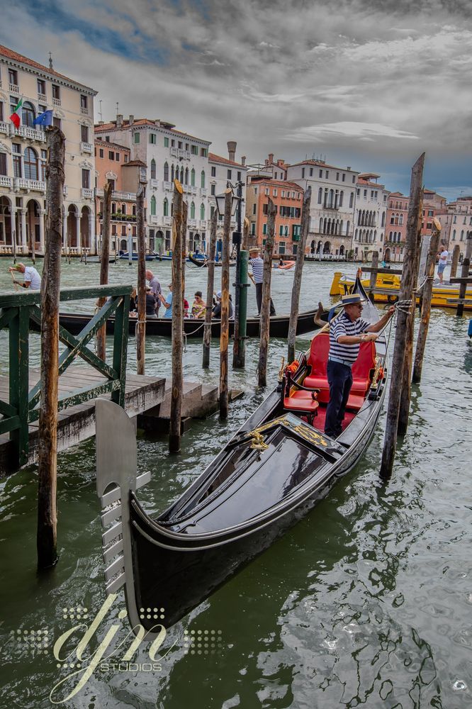 A gondolier ties up his gondola to a weathered wooden post that stands alongside a set of worn stairs. He awaits his next group of passengers with which to share the wonders of Venice.