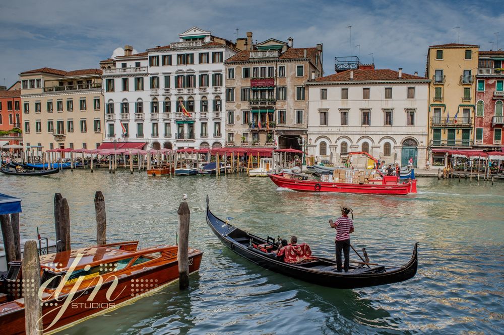 A gondolier looks off in the distance, down Venice's Grand Canal as he pilots his gondola. Tourists enjoy the ride past typical Venetian facades and other water crafts in the busy waterway. 