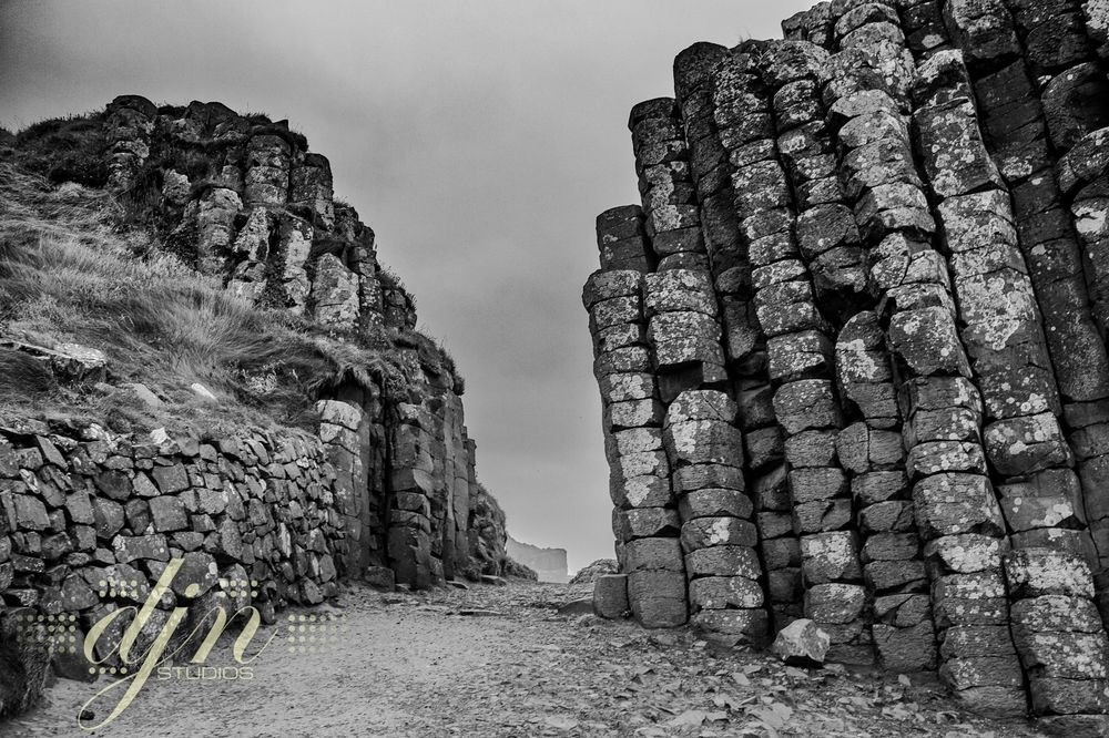 A black and white photo of a pathway through hexagonal shaped stone columns of basalt at Giants Causeway in Northern Ireland. Sea spray, and time have allowed the stone walls to grow a patina of lichen, moss and grasses. 
