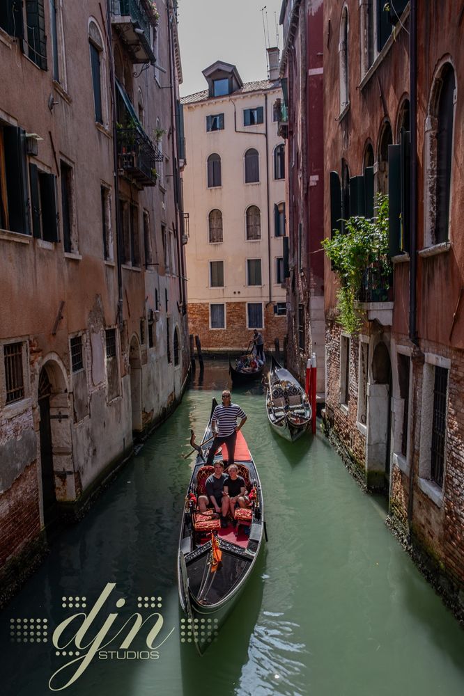 A gondola carrying two visitors, glides down a narrow canal in Venice, Italy. The boat approaches a small bridge after navigating the tight corridors of Venice's "roads". 