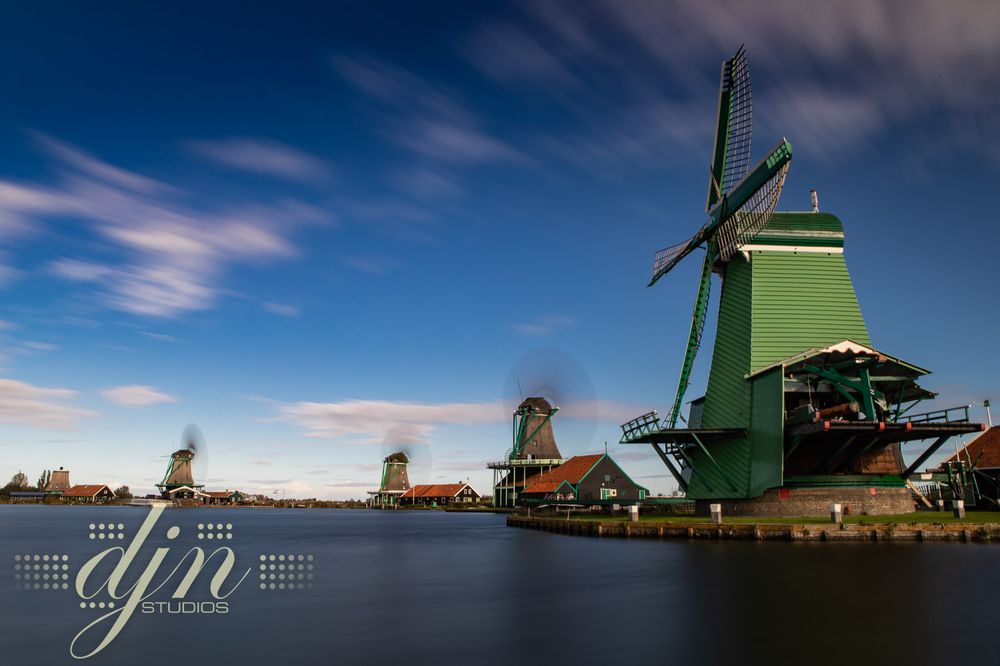 A long exposure photo of the 1600's era Dutch windmills in Zaanse Schans, along the water front. The windmill blades spin and blur into clean disks against the green wooden windmills structures, the clouds blur into long stretched white puffs in the sky. 