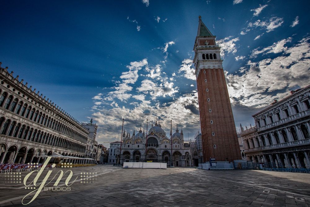 An early morning photo of Piazza San Marco in Venice, Italy. The Piazza is completely empty of the normal crowds of tourist. The camera faces the Basilica and the Campanile. The sky is half clear blue, and half clouds. Some of the clouds are thin and whispy while others are thick, dark and dense. Crepuscular light beams in around the thick clouds and through the thin ones. 