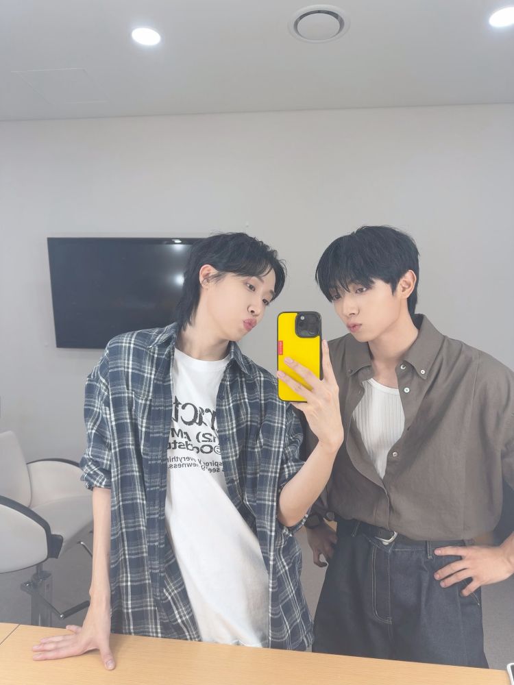 Dongmyeong anf Kang Hyungu from kband ONEWE. THey seem to be taking a mirror selfie, Dongmyeong leaning slightly on the table in front of them. Both are looking at the phone screen and pursing their lips, making duck faces.