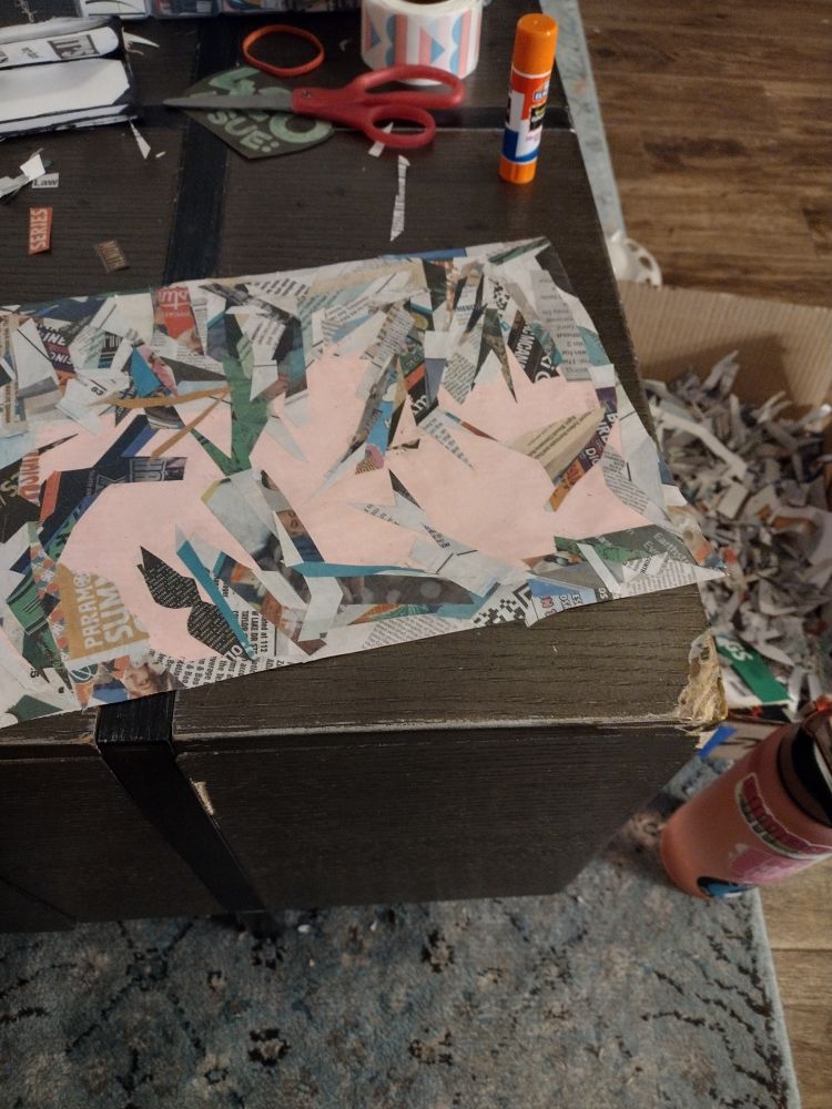 A picture of a work in progress on a brown coffee table. A pink piece of copy paper is mostly covered in triangular scraps of newspaper. A box of more scraps is next to the table.