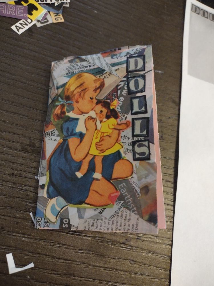 A picture of a mini zine sitting on a brown coffee table. The zine has a picture of a young girl in a blue dress kneeling and feeding a small doll in a yellow dress, with a background of newspaper scraps collaged together haphazardly. The word DOLLS is spelled out vertically in grey block letters.