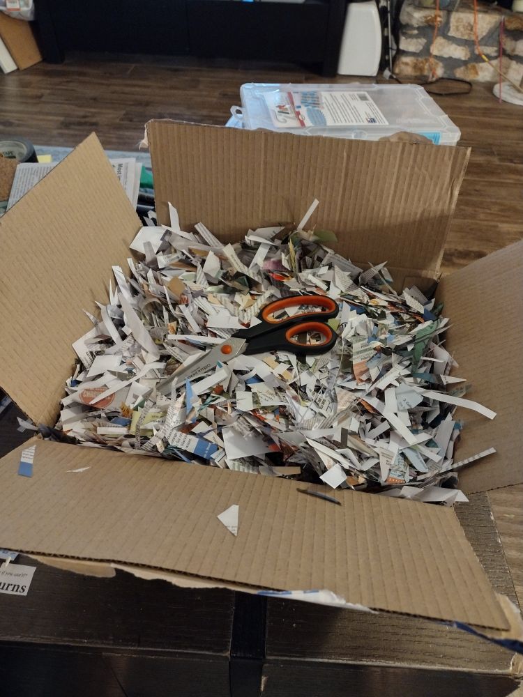 A picture of a cardboard box filled with scraps of paper of various sizes, shapes, and colors. A pair of large black and red scissors sit on top of the scraps