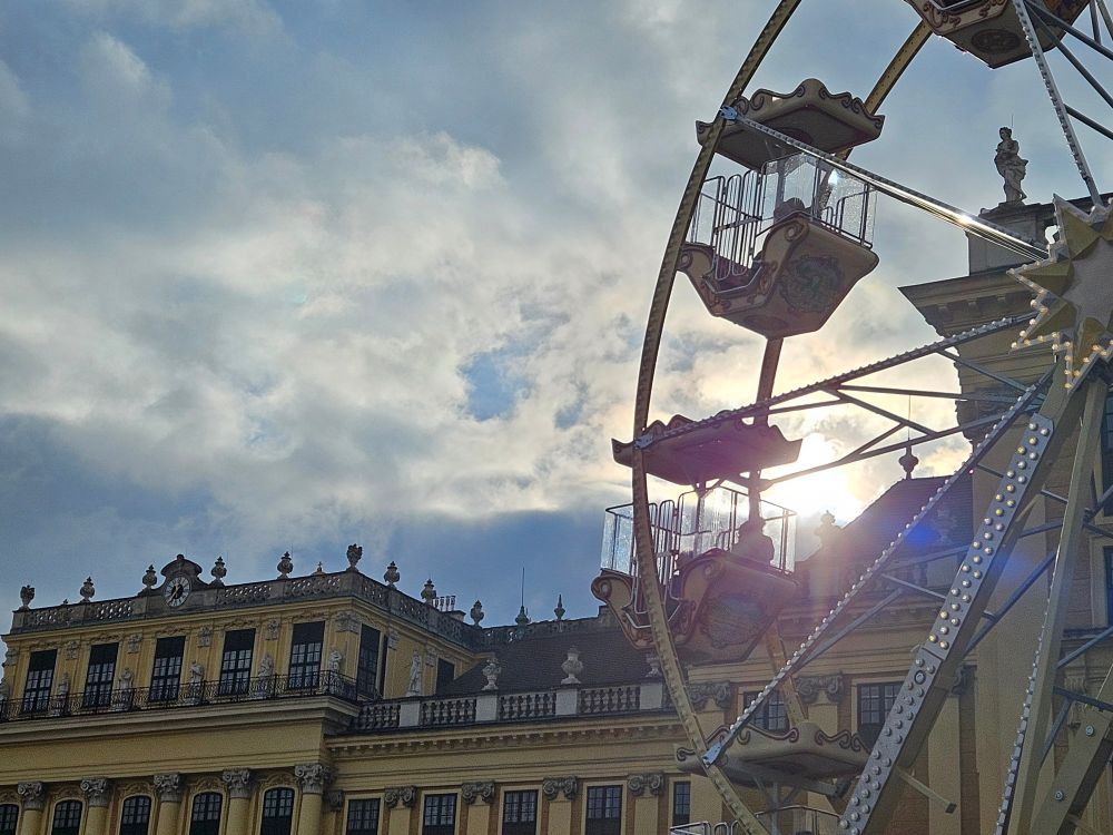 Bewolkter Winterhimmel, aber ein Sonnenstrahl blitzt hinter dem Schloss Schönbrunn hervor, davor ein vintage Riesenrad 