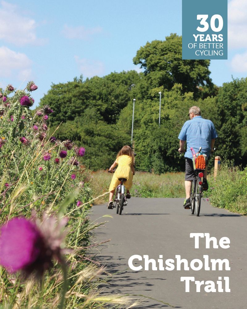 A girl in a yellow dress and a man in a blue shirt cycle along the Chisholm Trail. The wildflower verge to the left of the trail includes purple thistles. Overlaid is a '30 years of better cycling' logo and the text 'The Chisholm Trail'