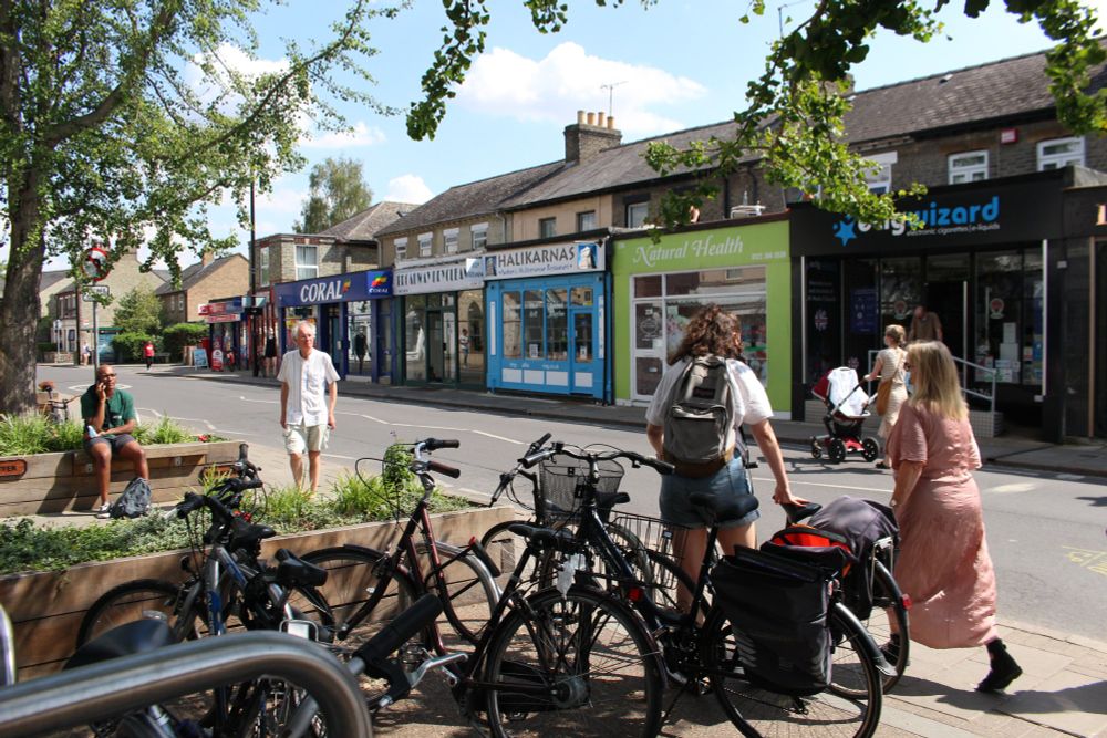 View of Mill Road from outside the Romsey Co-op showing people walking, wheeling a cycle and sitting on the public seating