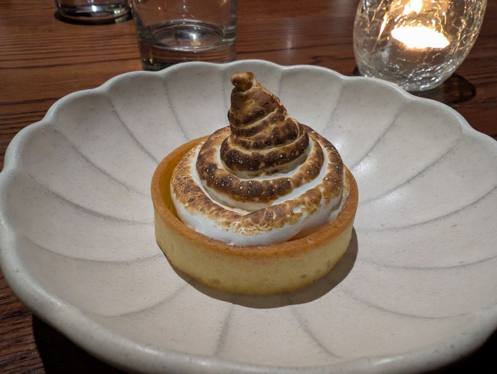 A Yuzu pudding from Sticks 'n' Sushi: A close-up shot of a lemon meringue tart with a golden-brown crust and a tall, swirled meringue topping, toasted to a light brown with darker brown highlights, sits in the center of a light-colored, fluted ceramic plate. In the background, a wooden table surface is visible, along with the base of a clear drinking glass and a lit candle in a glass holder, casting a warm glow.