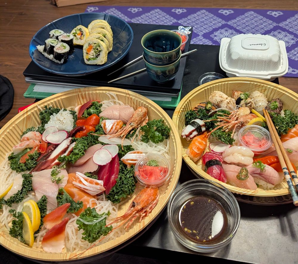 A small coffee table with two platters of sashimi from a wide variety of sea creatures, a plate with rolls, a container of edamame, and two small cups waiting for the sake to heat. 