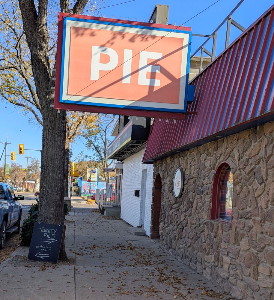 A single story building with a stone facade and a red roof. A large sign hangs over the sidewalk that just says PIE in white letters on red. A chalk sandwichboard is leaning up against a tree next to the road with an arrow pointing towards the business saying "Turkey Pies" in large friendly letters, and another arrow pointing towards the street labelled with sad letters reading "no turkey pies." 