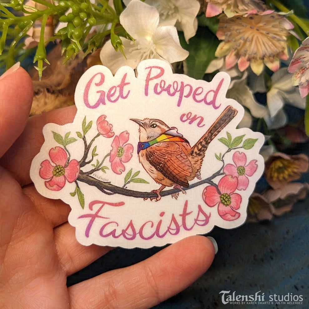 My hand holding a sticker in front of some fake flowers. The sticker is my art of a grumpy Carolina  wren on a flowering dogwood branch. The text reads: "get pooped on, fascists." The wren is also  wearing a progress pride cloak.