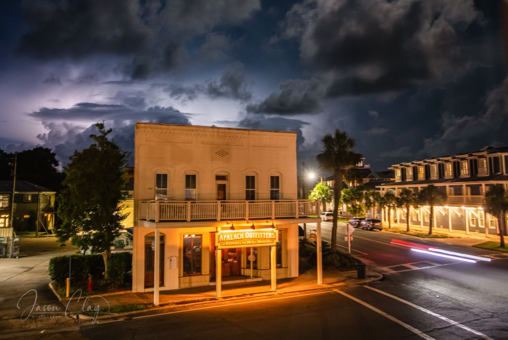 A nighttime image of a building in Apalachicola, Florida with heat lightning striking in the background. In the foreground, long streaks of a car's headlights reflect on the asphalt. 