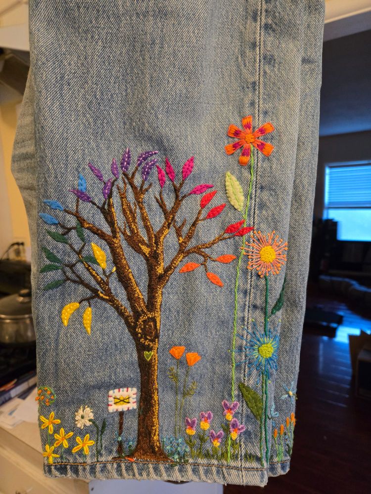 Hand embroidery on Levi's 501 jeans featuring a tree made of different color french knots with rainbow colored felt applique leaves. Miscellaneous flowers, two California poppies, and a No Kings protest sign are included in the scene. 