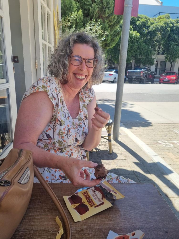 A curly haired white person wearing glasses and sitting outside a shop eating the chocolate double fudge brownie icecream sandwich and laughing because it's so good