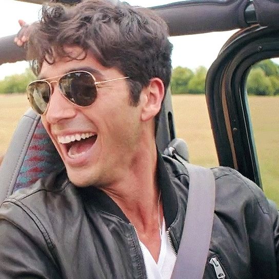 A dark-haired man laughing while wearing a black leather jacket over a white T-shirt and aviator sunglasses