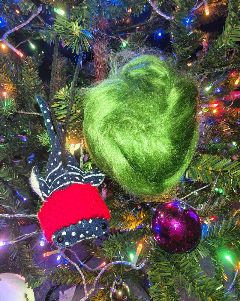 a bundle of bright green wool, roughly the colour of brussel sprouts, lies nestled in the same tree but with different lighting. there's a fabric ornament shaped like a whale shark wearing a knitted red scarf
