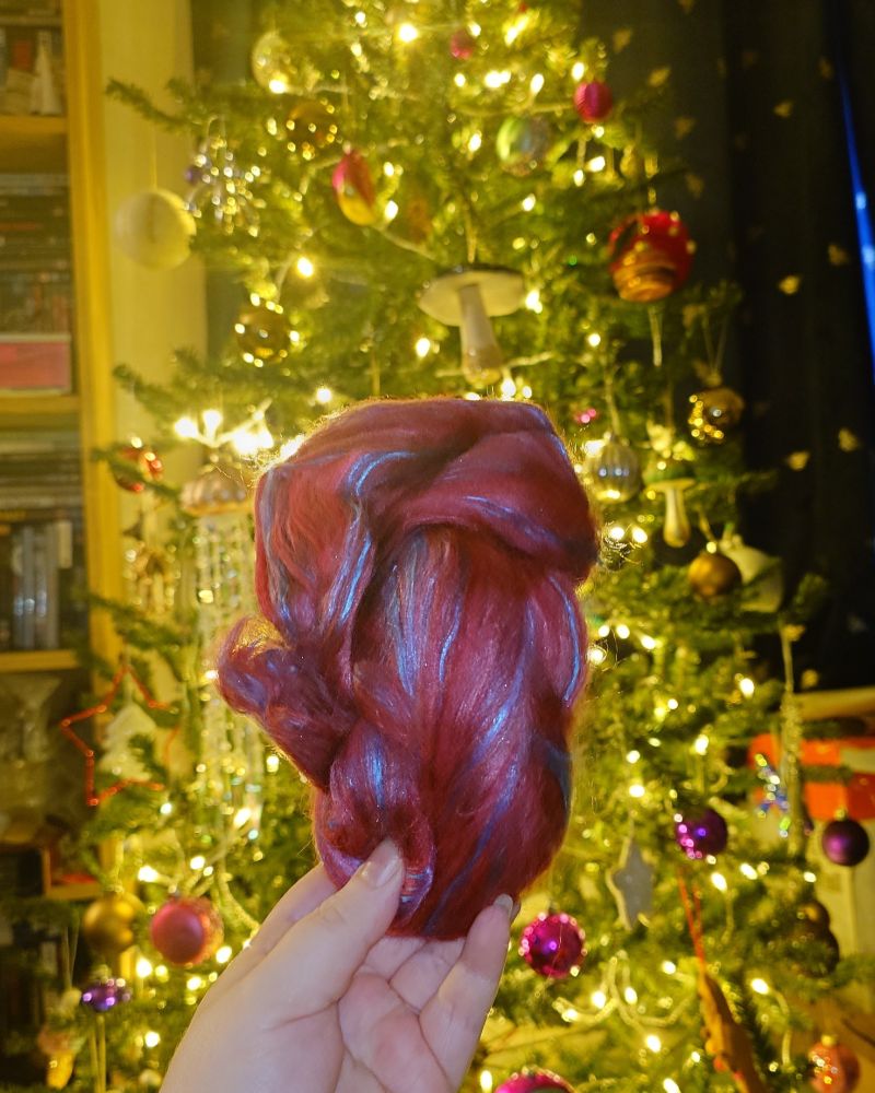 my hand holds the bundle of fibre in front of the tree, the lights are warm white here and the fibre is a deep red with ribbons of a shiny blue running through it