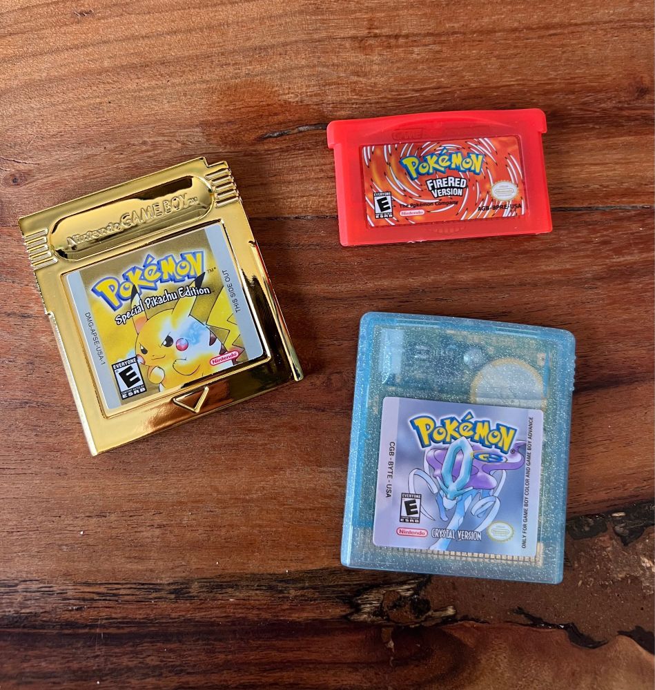 Pokémon yellow, Pokémon Firered, and Pokémon crystal gameboy games with new game stickers on them. The Pokémon yellow game is in a gold casing.