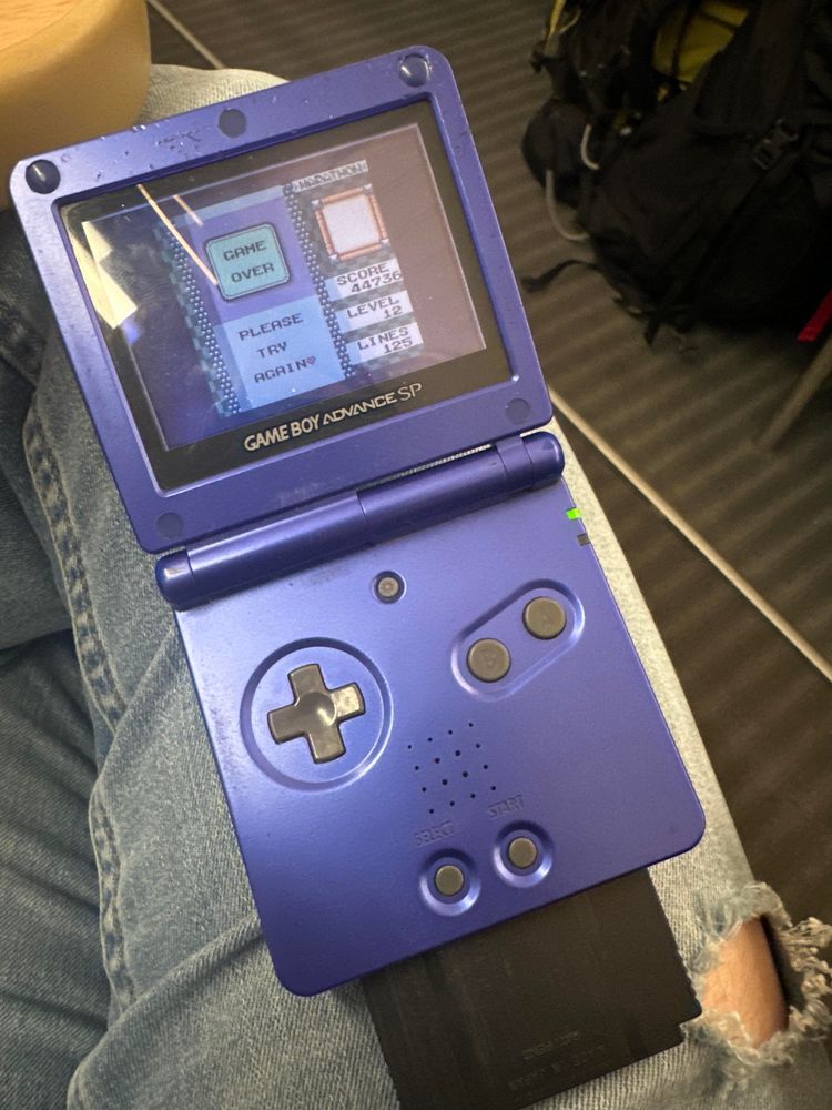 An open purple Game Boy Advance SP is displayed. The screen shows a "Game Over" from Tetrix DX message with a score and options to try again. A game cartridge is inserted at the bottom.