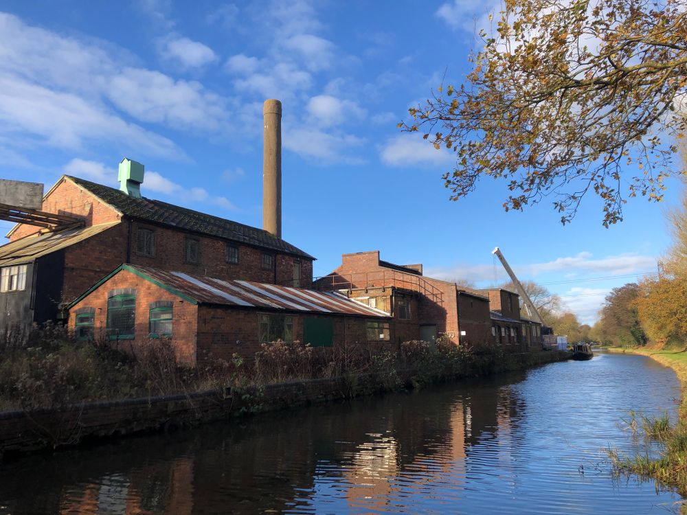 Industrial buildings from the 19th century on the left with canal running past to centre and right. Buildings include a chimney, warehouse and workshop.