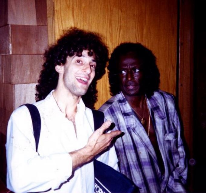 A picture of a young, backpack wearing Kenny G, smiling and pointing at iconic jazz legend Miles Davis, who looks at Kenny with deep dismay and concern, as if he thinks something is wrong with the excited soprano saxophonist.