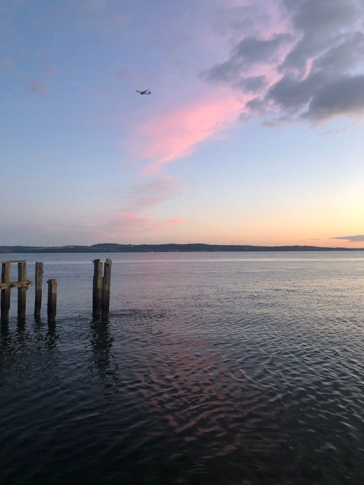 Sea with some old wooden uprights and land on the horizon. Pink cloud and itโs reflection, aeroplane