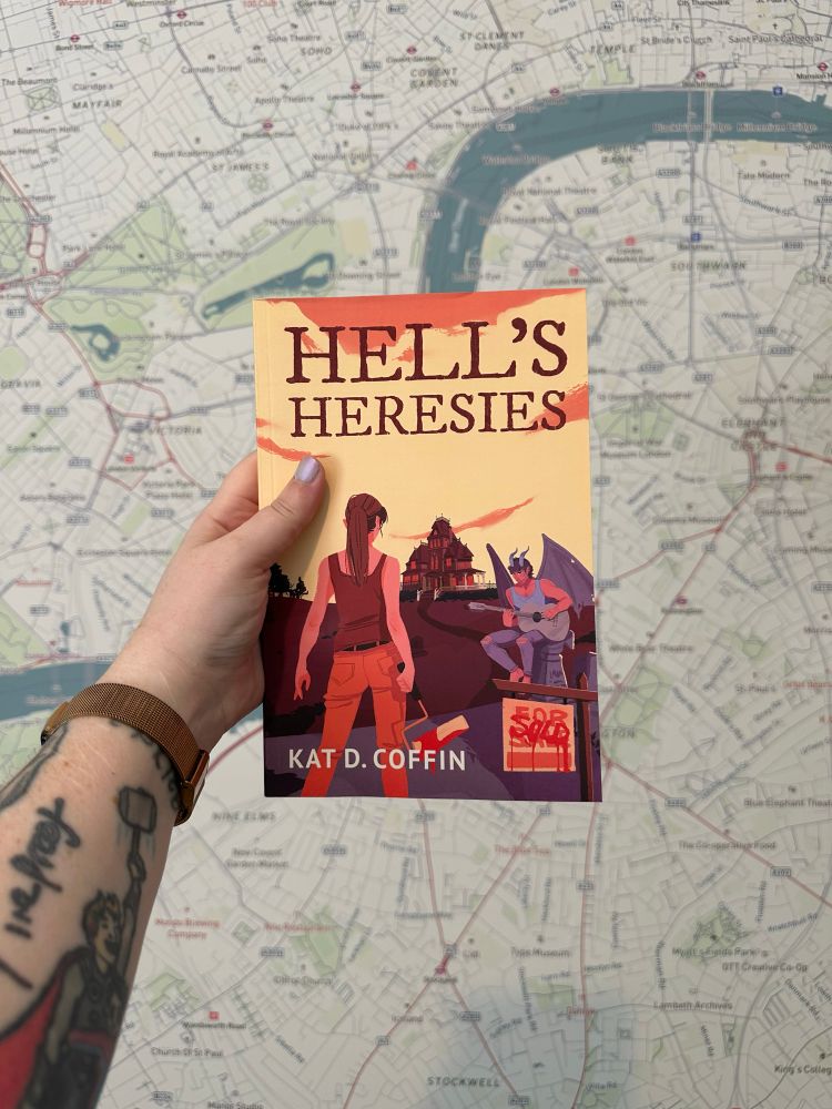 a copy of Hell’s Heresies by Kat D. coffin held up against a wallpapered background of London, England
