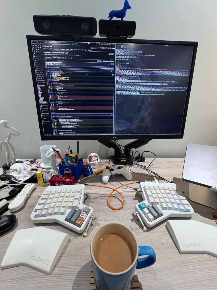 my desk with my morning coffee, split keyboard, and my monitor with GitHub and Ghostty terminal open. There is a collection of clutter below and to the left of the monitor 