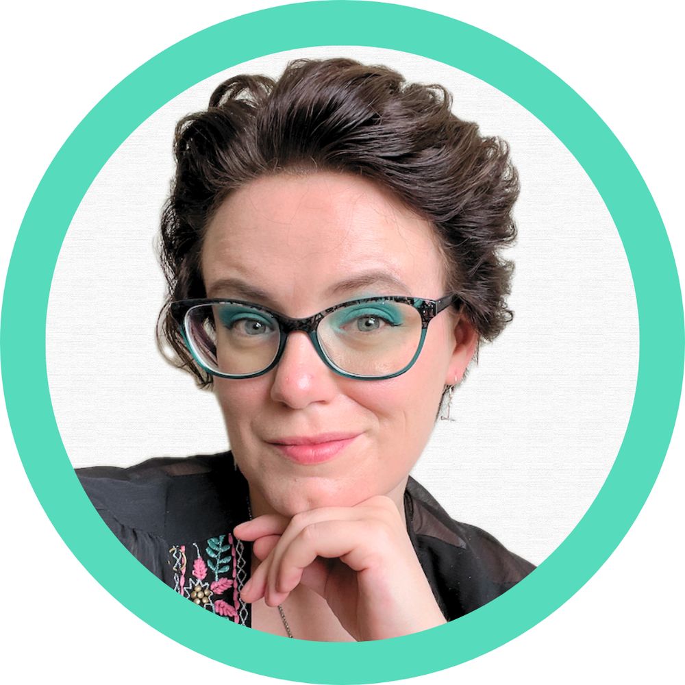 Headshot of Mel Lee-Smith, freelance writer and social justice advocate. She has short, black curly hair. She's wearing blue eyeshadow with blue and black glasses, as well as a black sheer blouse with blue and pink embroidered flowers. Her chin is resting on her left hand.