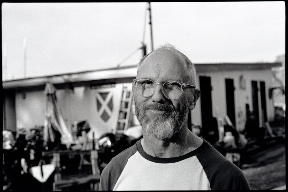 Black and white photograph. A man stands in front of the camera looking mostly for unimpressed withe the hual out situation. 