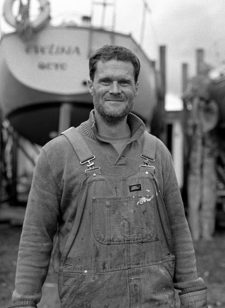 Black and white photograph. A man stands looking into the camera wearing stained overalls and an old knitted sweater. 