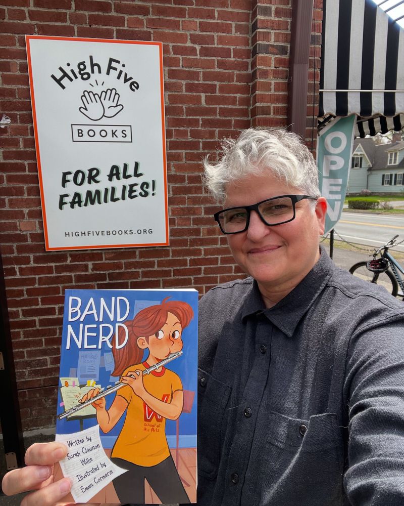 Me - person w shirt gray hair and black glasses, standing in front of High Five Books store sign, holding a copy of Band Nerd.