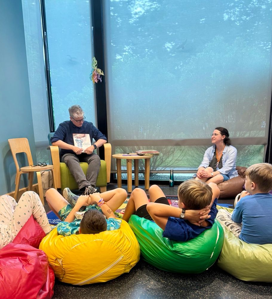 Me reading Molly’s Tuxedo to a gathering of children sitting on colorful bean bag chairs. Librarian sits next to me watching. 