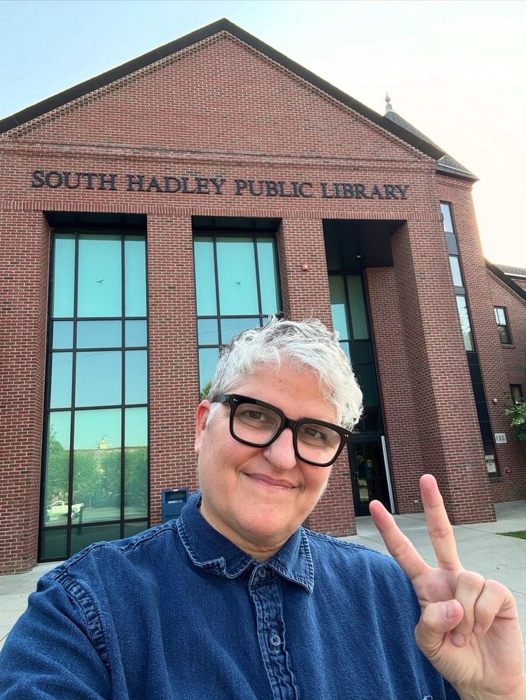 Gray haired me w black glasses and blue shirt flashing a peace sign out front of the library